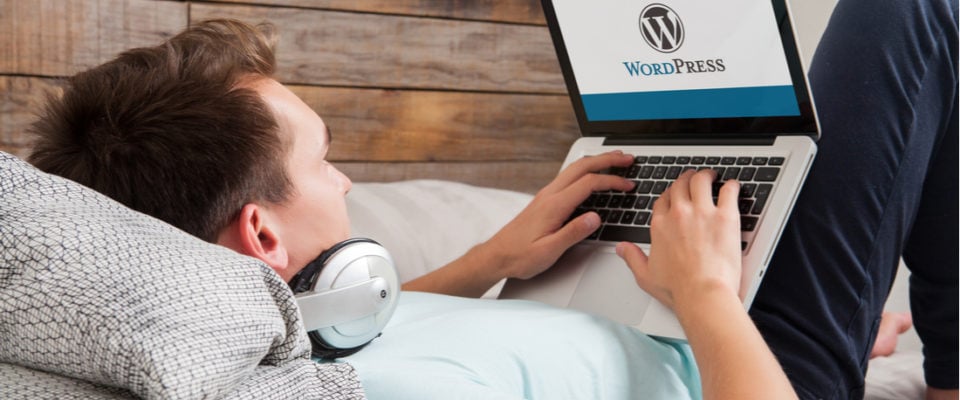 Photo of man lounging in bed using laptop to access WordPress with headphones