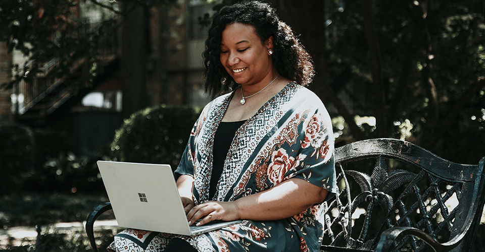 a Black woman sits on an outdoor bench on a sunny day while working at her laptop and smiling