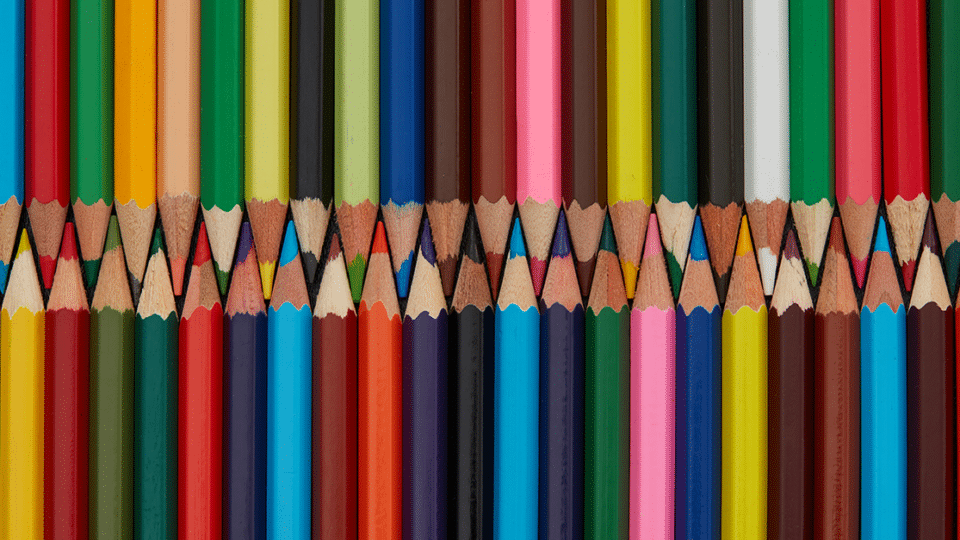 several colored pencils laid out on a table. The sharpened ends are fitted together as the top line of pencils points down and the bottom line of pencils point up