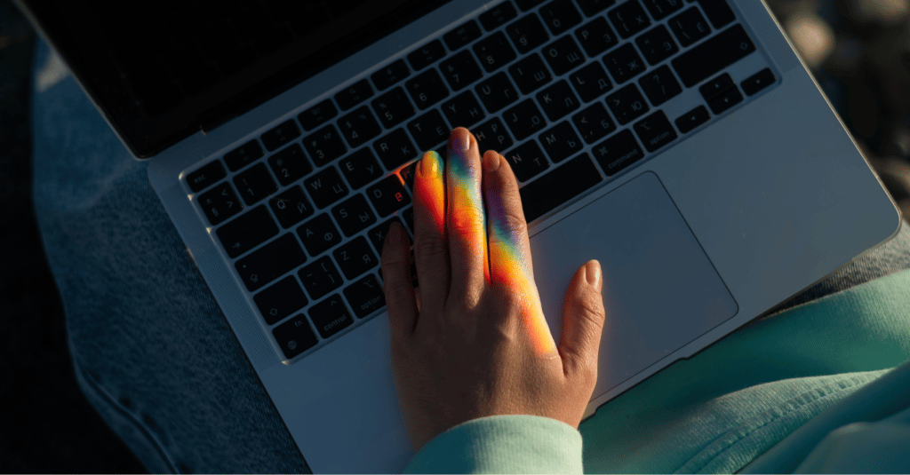 a person's hand hovers over a laptop keyboard. a rainbow prism falls over their fingertips
